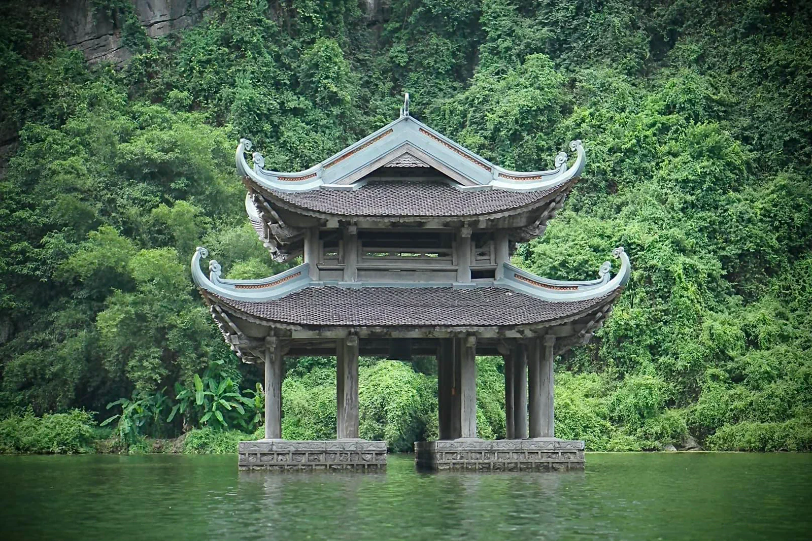 Two-tier traditional pavilion standing in the water below forested limestone cliffs at Trang An